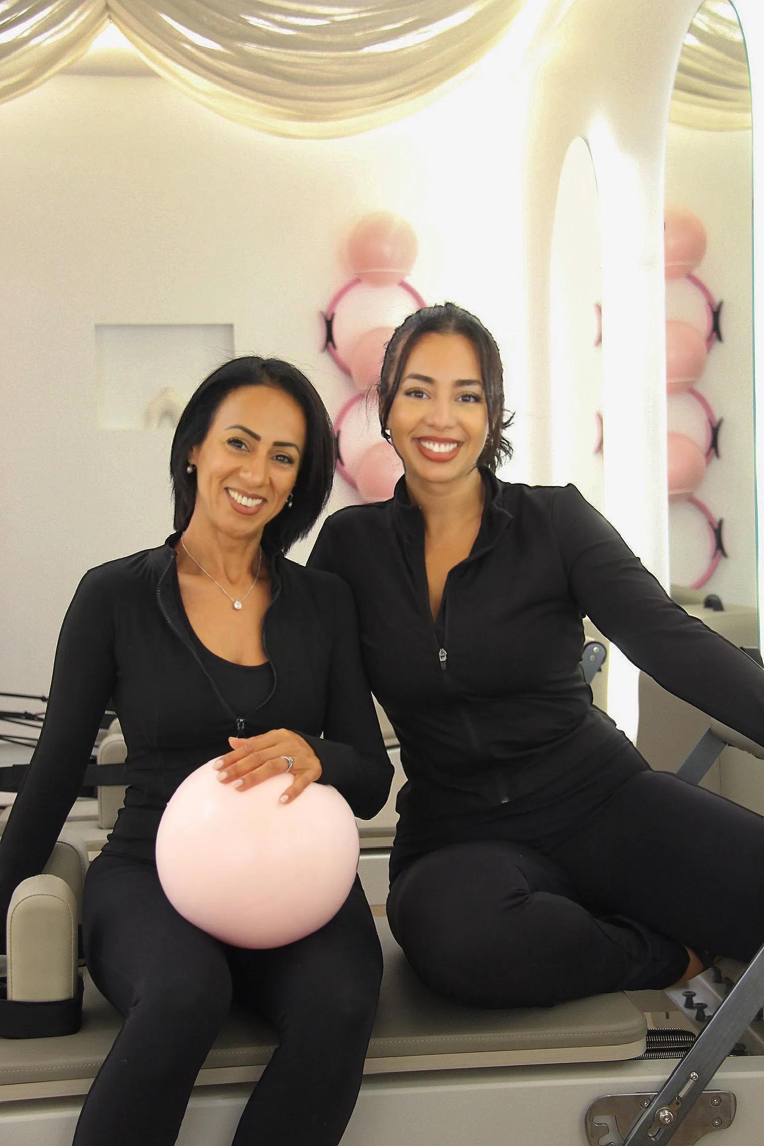 Les deux fondatrices du studio, souriantes, assises sur un reformer avec un ballon rose